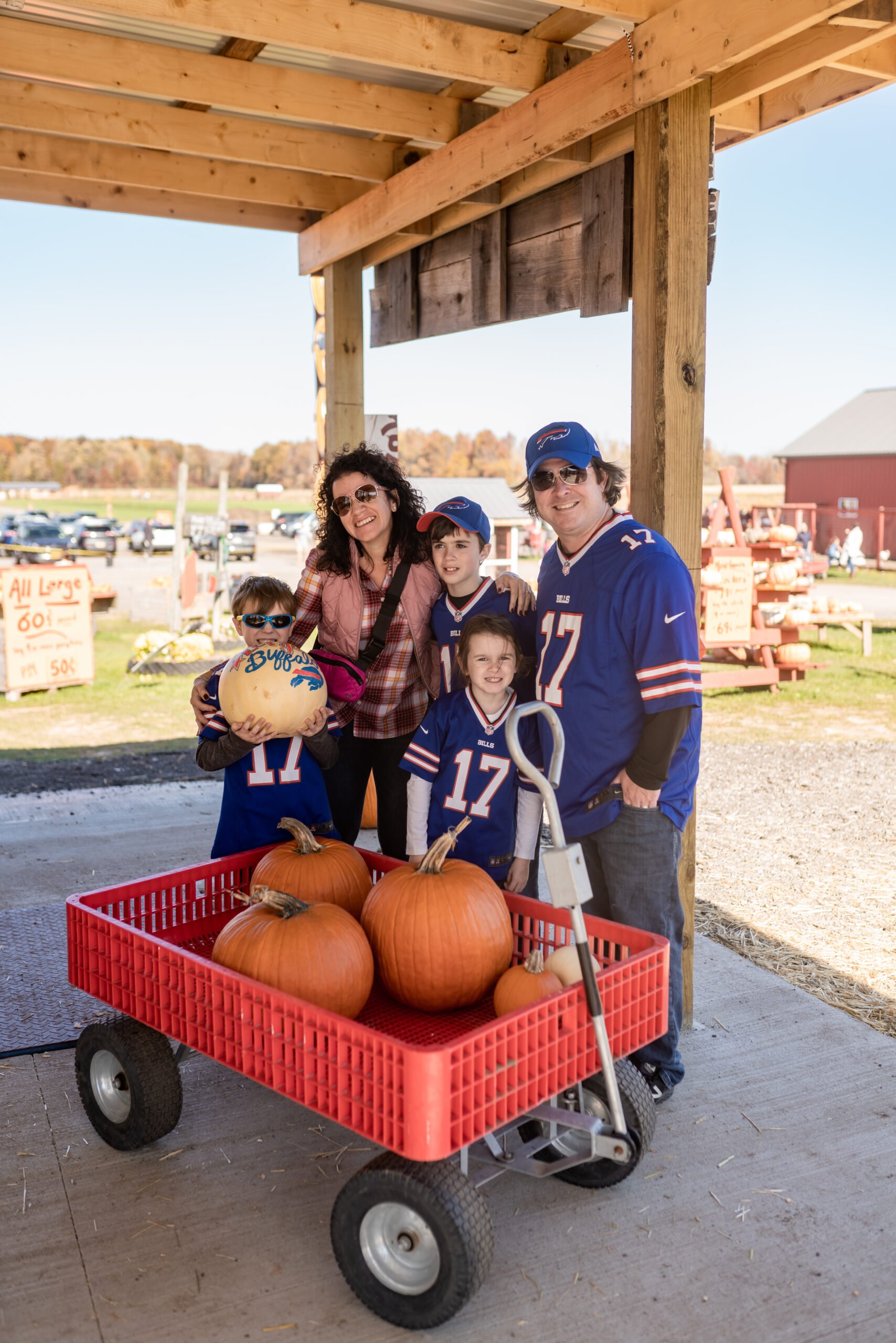 Family enjoying a fall day at a pumpkin patch, posing with a red cart filled with pumpkins, wearing matching jerseys, in a sunny outdoor setting.