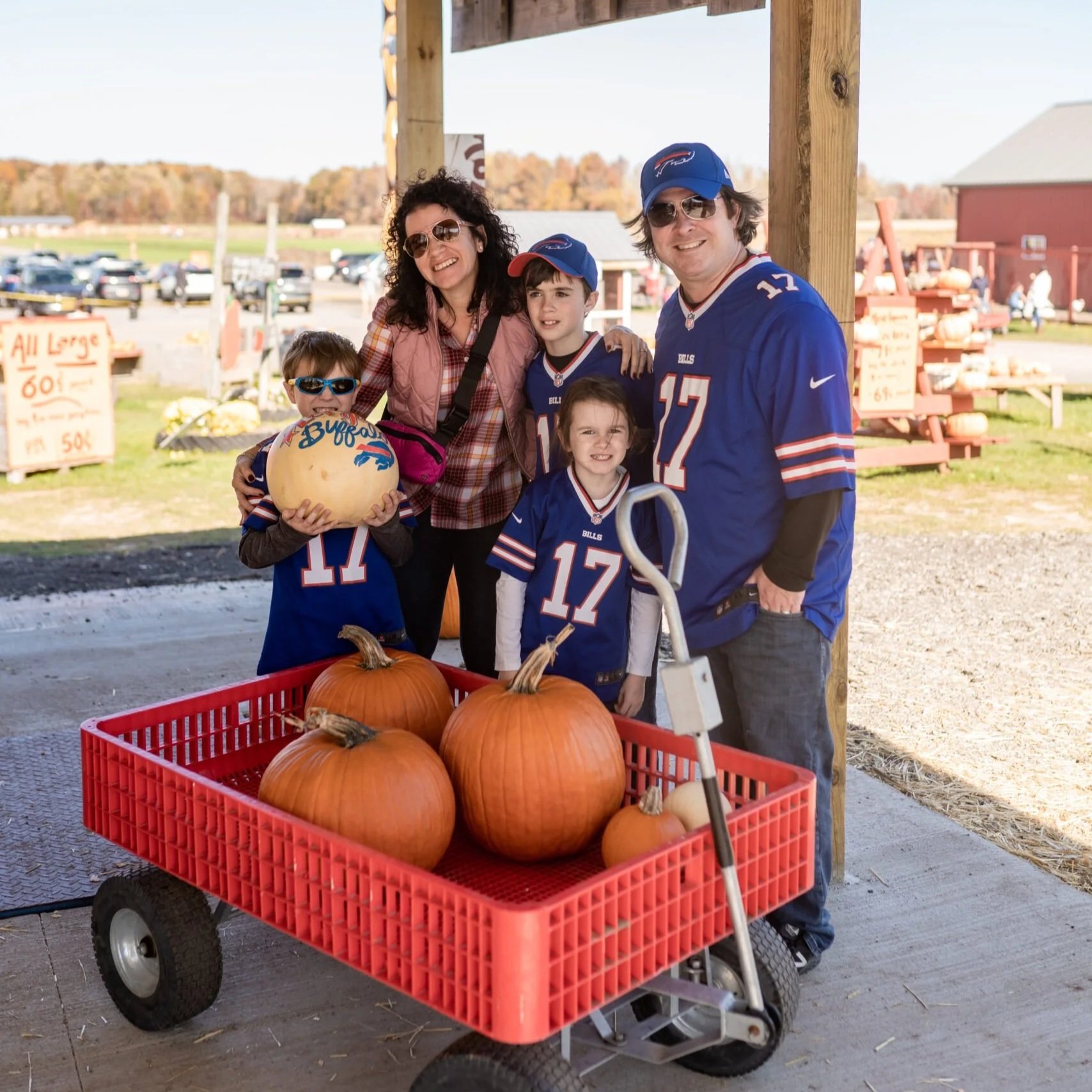 Family enjoying a day at a pumpkin patch, wearing Buffalo Bills jerseys, posing with a cart filled with pumpkins and a decorated gourd. Bright autumn scenery in the background.