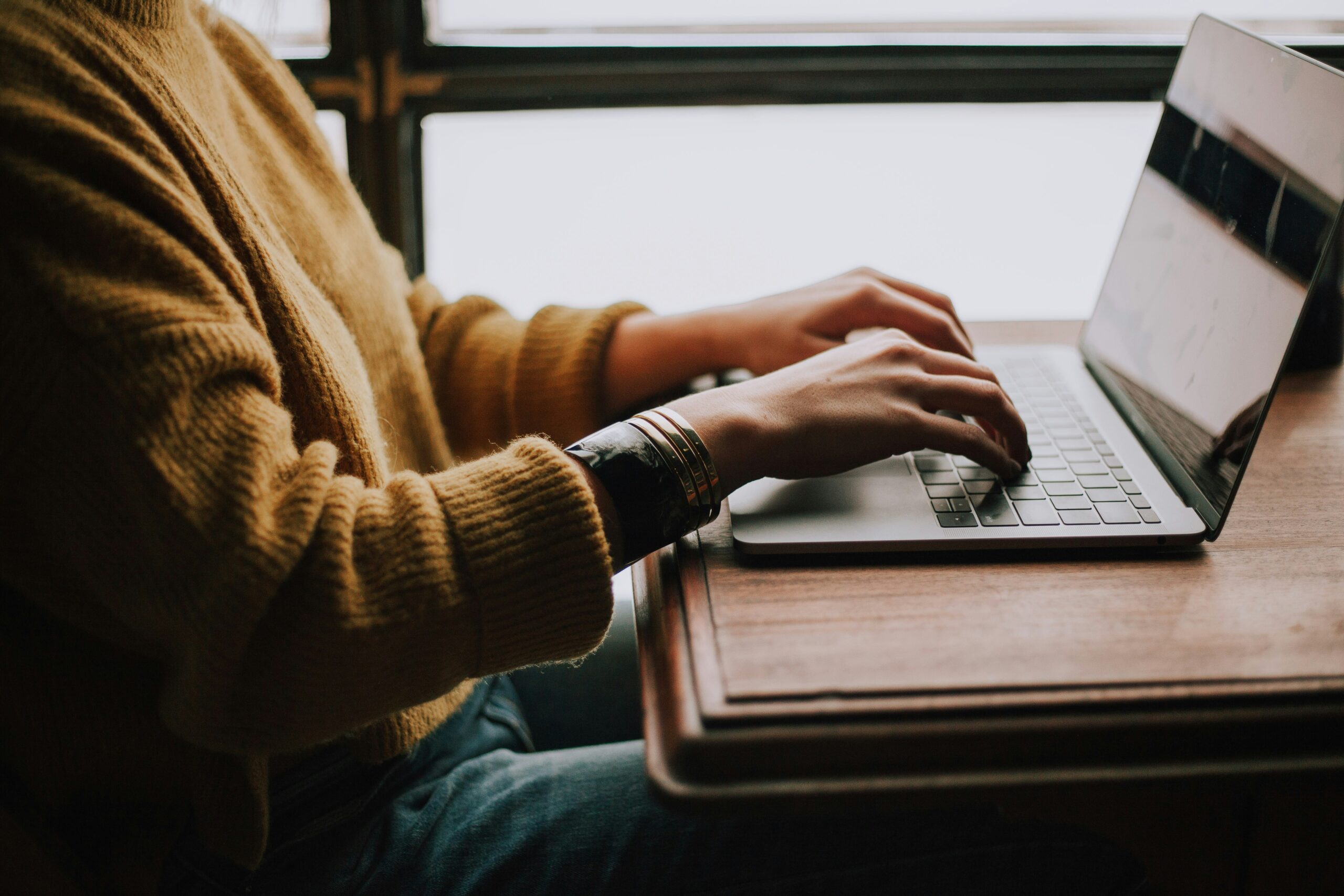 christin-hume-mfB1B1s4sMc-unsplash Person typing on a laptop while sitting at a wooden table, wearing a cozy yellow sweater, in a bright indoor setting with natural light.