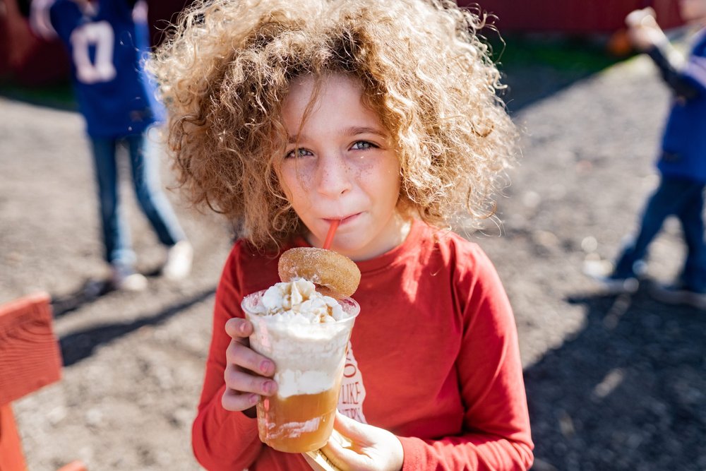 Child enjoying a delicious dessert drink topped with whipped cream and a donut, wearing a red long-sleeve shirt, with a playful outdoor setting in the background.