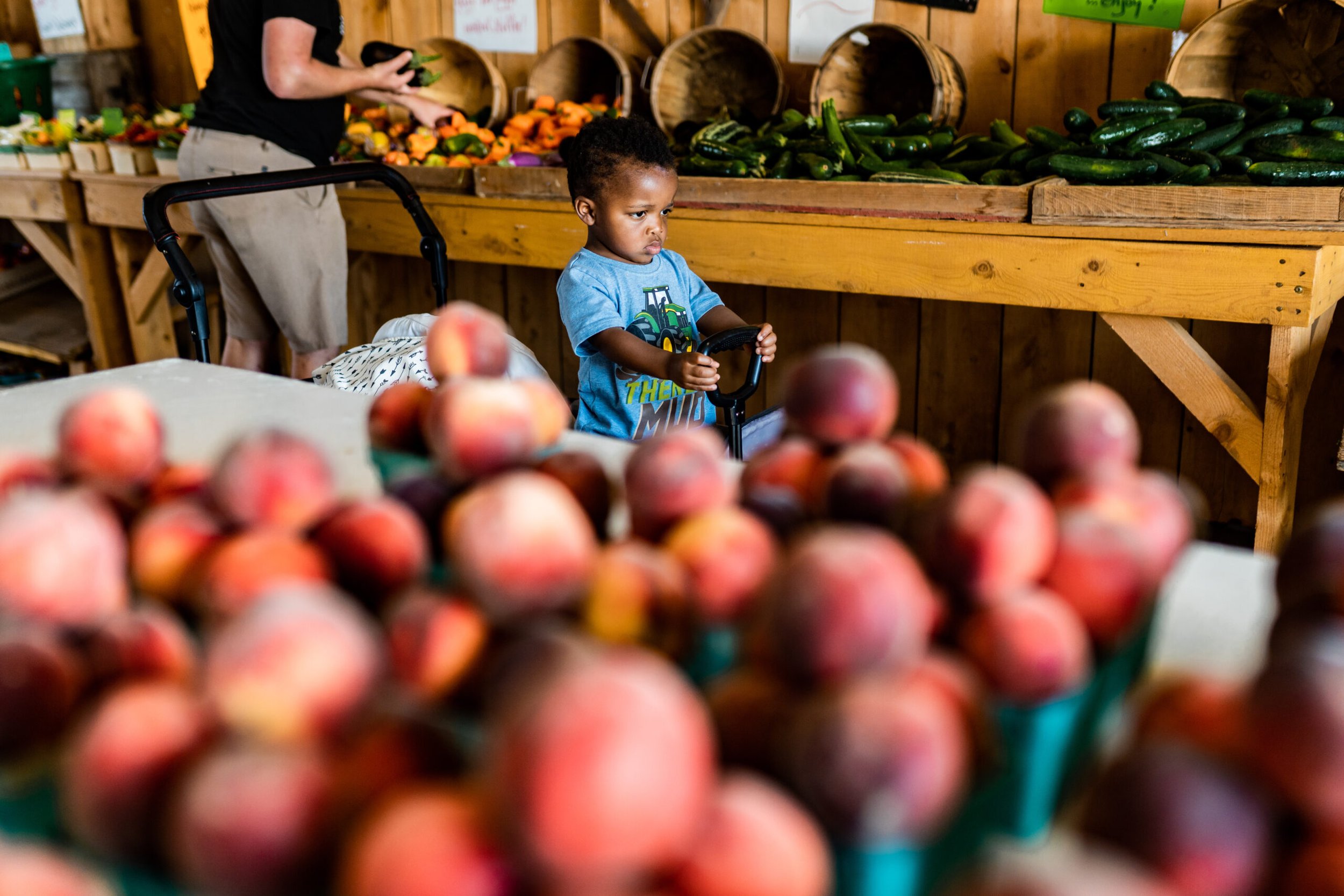 Child exploring a local farmers market, surrounded by fresh peaches and vegetables, while an adult shops in the background.