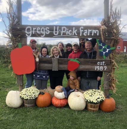 Group of people posing at Greg's U-Pick Farm, established in 1987, surrounded by pumpkins and decorative flowers, with a sign featuring a red pepper and a strawberry. The image captures the essence of a family-friendly farm experience during the fall season.
