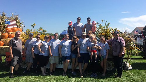 Group of people posing together in front of a decorated trailer with pumpkins and corn, celebrating a fall harvest festival on a sunny day.