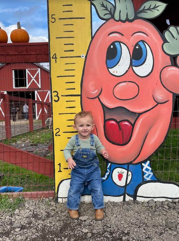 A happy toddler in denim overalls stands next to a colorful height chart featuring a cartoon carrot with a smiling face at a farm, surrounded by pumpkins and a barn in the background.