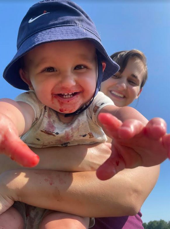 Smiling baby in a blue bucket hat reaching out with hands covered in red stains, held by a woman with short hair, under a clear blue sky. The scene captures a joyful outdoor moment, highlighting the playful interaction between the child and the caregiver.
