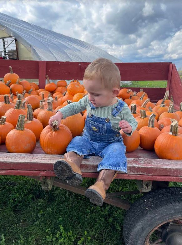 Toddler in denim overalls sitting on a wagon filled with vibrant orange pumpkins at a pumpkin patch, with a cloudy sky in the background.