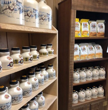 img-0199 (1) Shelves displaying various bottles of pure maple syrup and packages of pancake mix in a rustic store setting, highlighting local food products.