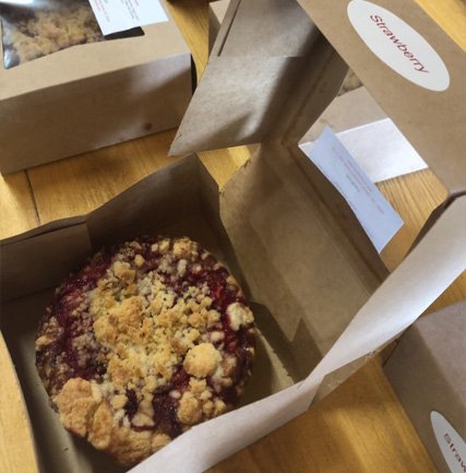 img-0203 (1) Freshly baked strawberry crumble cake in an open cardboard takeout box, placed on a wooden table, with additional dessert boxes visible in the background.