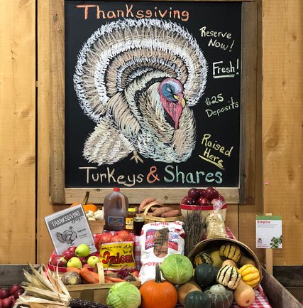 Colorful chalkboard sign promoting Thanksgiving turkeys with a detailed turkey illustration, featuring text about reserving fresh turkeys with $25 deposits. Surrounding the sign are fresh fruits, vegetables, and other seasonal produce, creating a festive display.