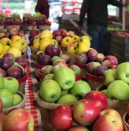 Baskets filled with a variety of fresh apples, including green, red, and yellow, displayed at a fruit market with people in the background.