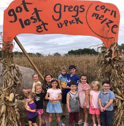 A group of children poses in front of a colorful sign that reads "Got Lost at Greg's U-Pick Corn Maze," surrounded by tall corn stalks in a farm setting.