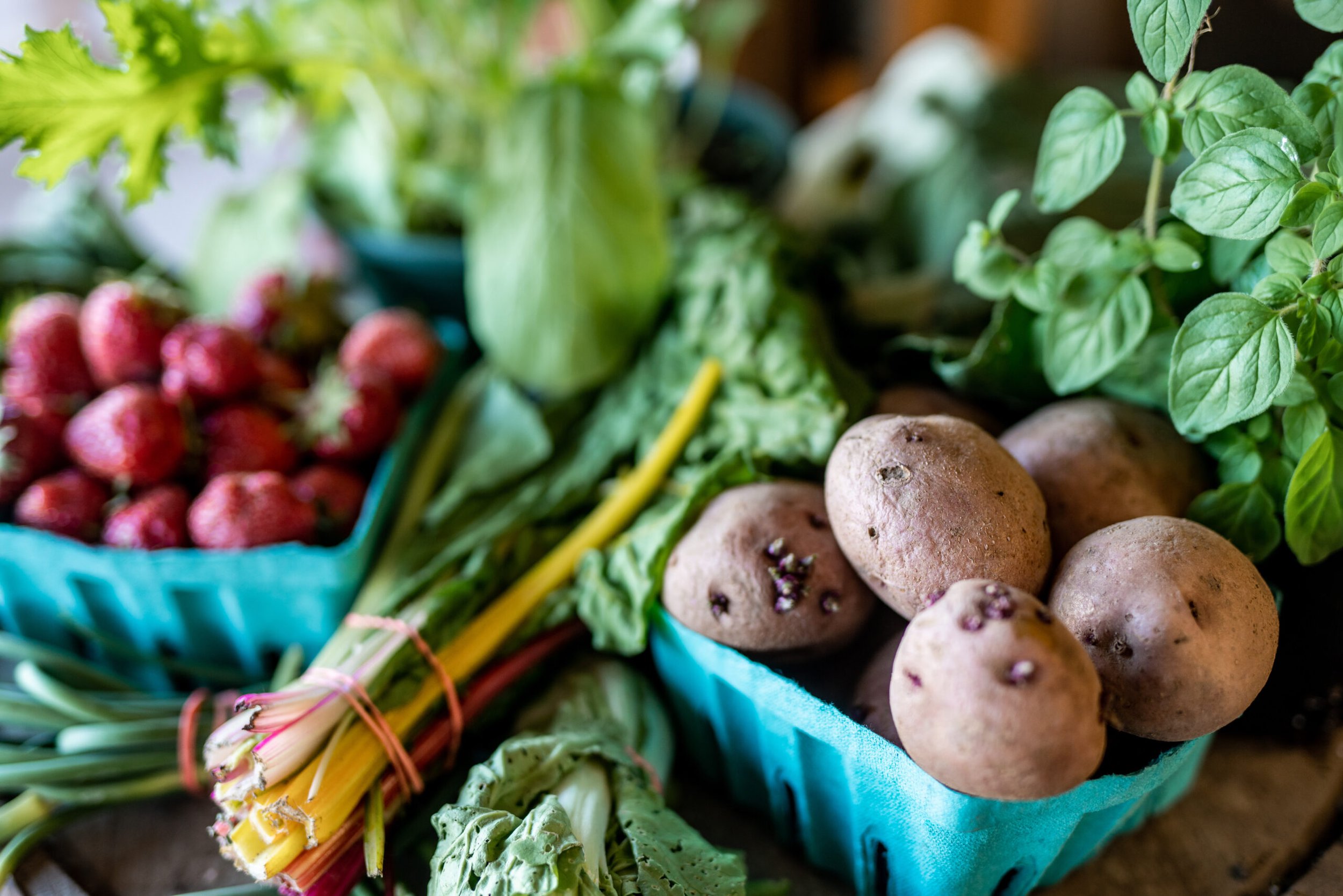 Fresh produce assortment featuring strawberries, leafy greens, and potatoes in blue baskets, highlighting seasonal vegetables and healthy eating options.