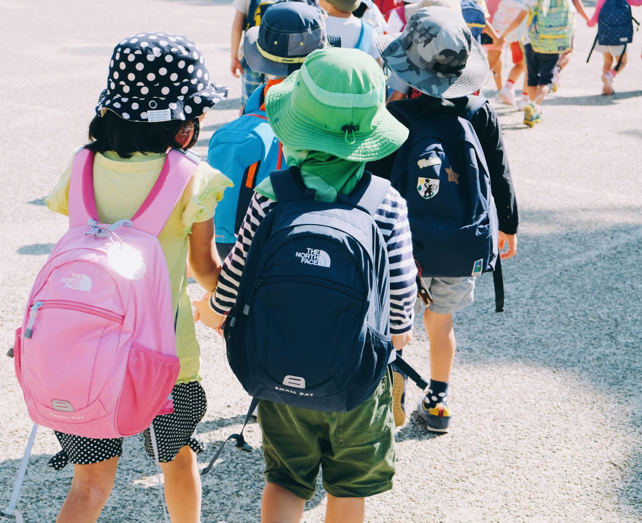 Children walking together outdoors, wearing colorful backpacks and sun hats, enjoying a sunny day.