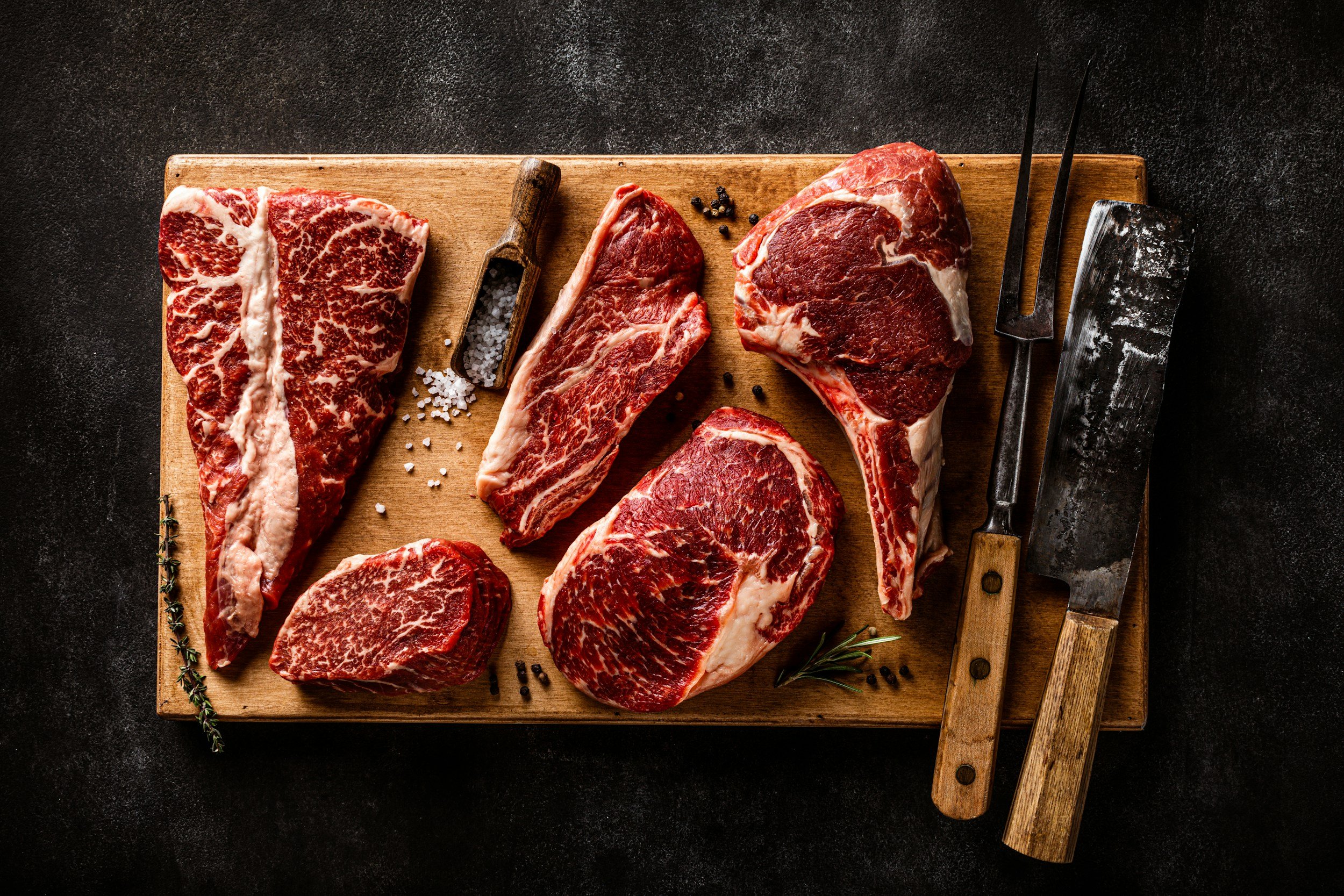 unsplash-image-cEJGctX_DgI (1) Assorted cuts of premium beef, including ribeye and T-bone steaks, displayed on a wooden cutting board with seasoning and butchering tools. Ideal for culinary enthusiasts and meat lovers.