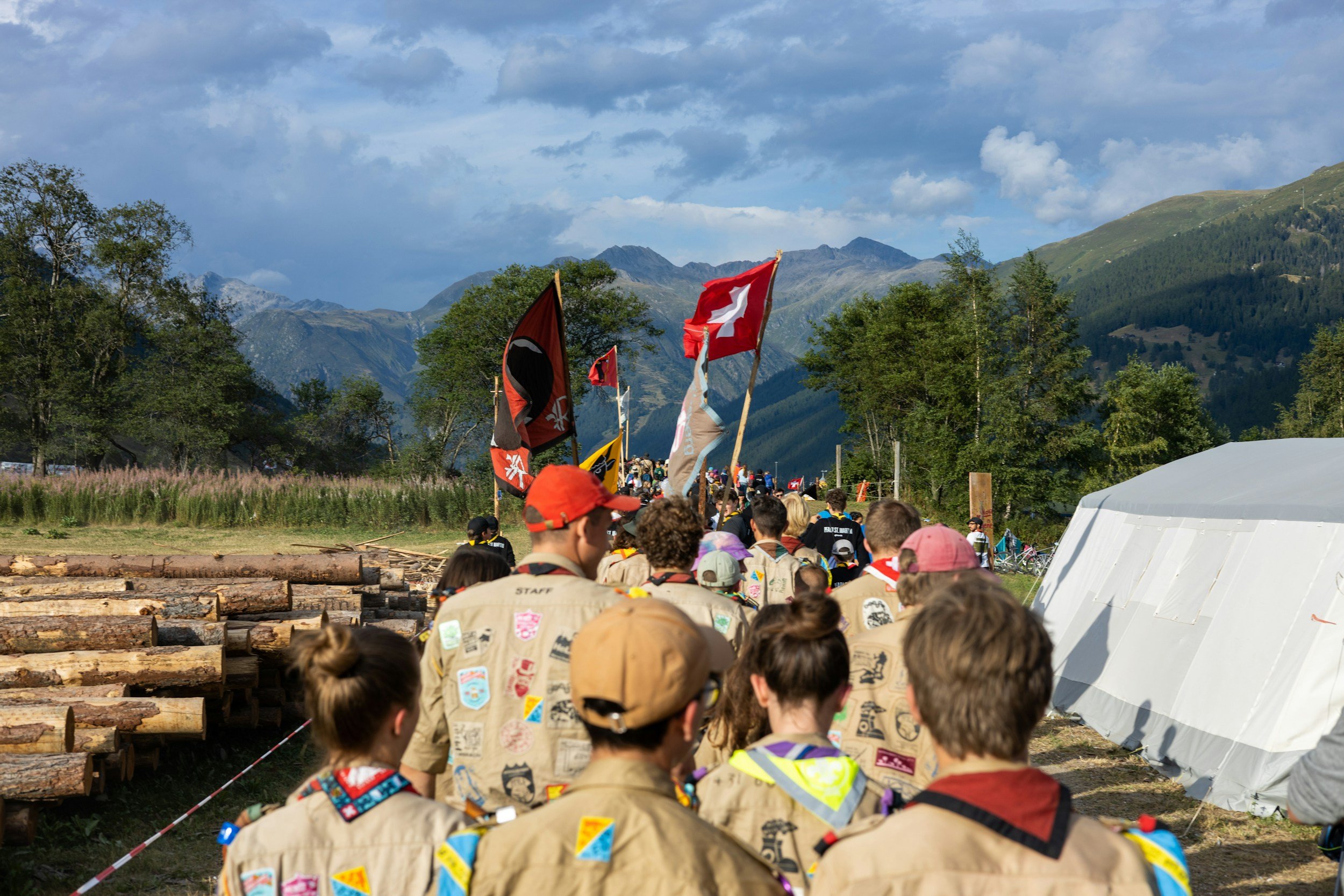 A group of scouts wearing uniforms with various badges walks along a path in a mountainous area, carrying flags, with logs stacked nearby and a tent visible in the background. The scene captures a vibrant outdoor activity, showcasing teamwork and camaraderie among participants.